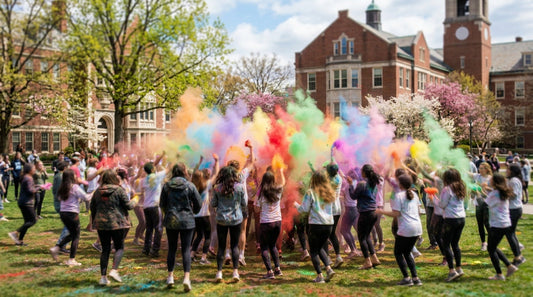 Students seen from behind throw clouds of colorful powder on a sunny university quad.
