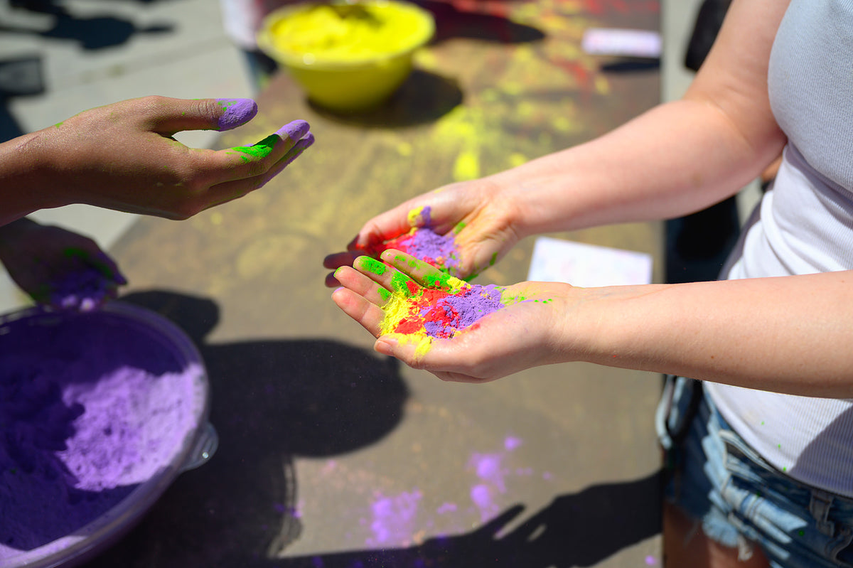 Hands holding red, yellow, green and purple cornstarch color powder over a bowl of purple powder